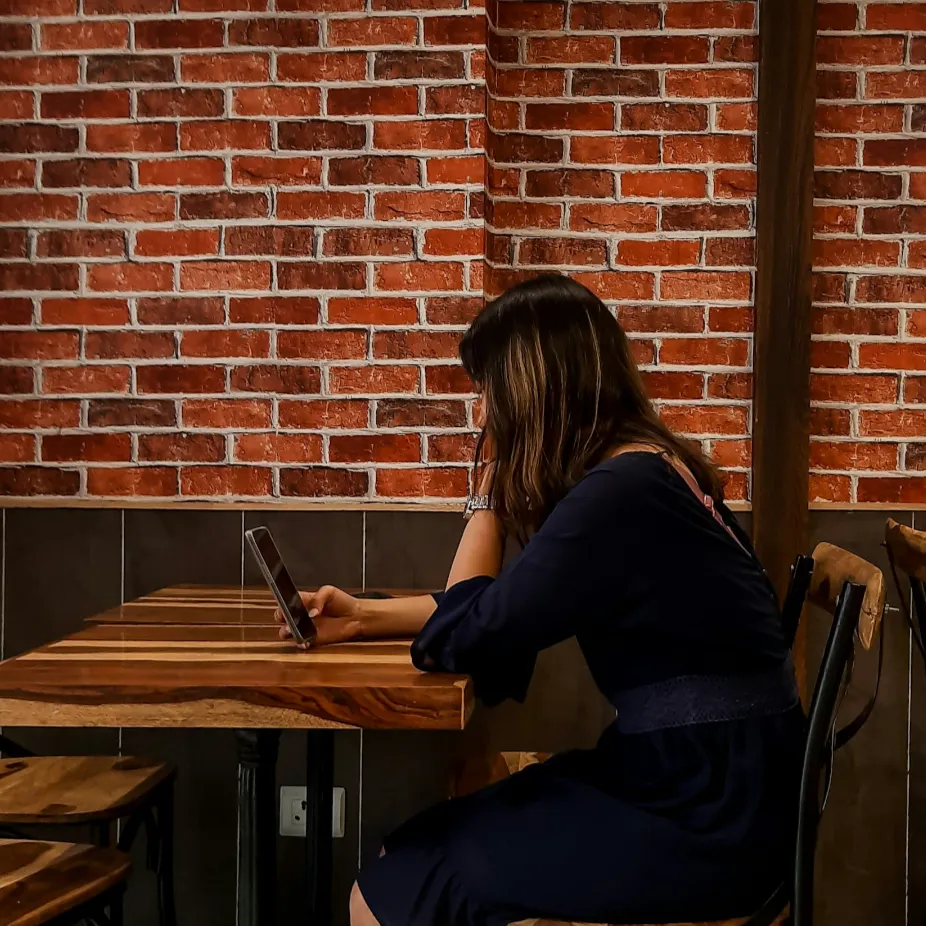 A woman in a dark dress sits alone at a wooden table in a cafe, looking at her phone. The wall behind her has a red brick pattern, creating a cozy atmosphere.