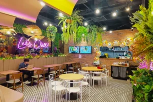 Cozy café with brick walls, hanging plants, and a neon "Let's Chill" sign. A person works on a laptop at a table, creating a relaxed vibe.