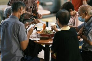 A group of six older adults gather around a round table, joyfully sharing a meal with chopsticks. They appear engaged and content, fostering a warm, communal atmosphere.