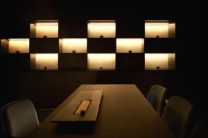 Dimly lit room with a minimalistic design. Illuminated shelves display small objects, casting a warm glow. A modern table and chairs are in the foreground.