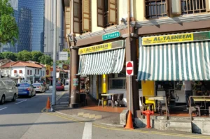 Street corner with Al-Tasneem restaurant, featuring green-striped awnings. Signs indicate "Jln Pinang" road. Cars and trees are visible under a clear sky.