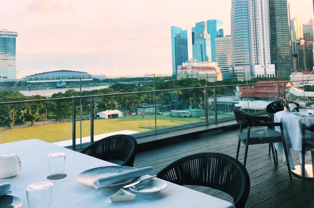 Rooftop terrace with tables set for dining, offering a panoramic view of a city's skyline. Green space and a large, modern building are visible. Calm and inviting ambiance.