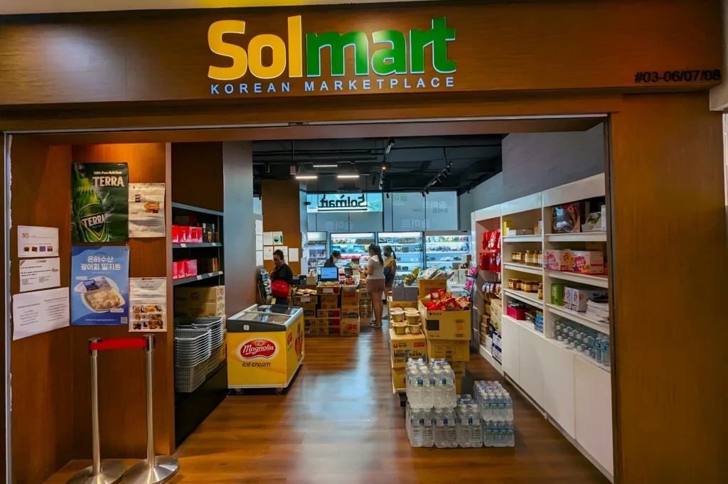 Entrance of Solmart Korean Marketplace, showcasing aisles with Korean products. Two shoppers explore near the checkout counter. Warm, inviting ambiance.