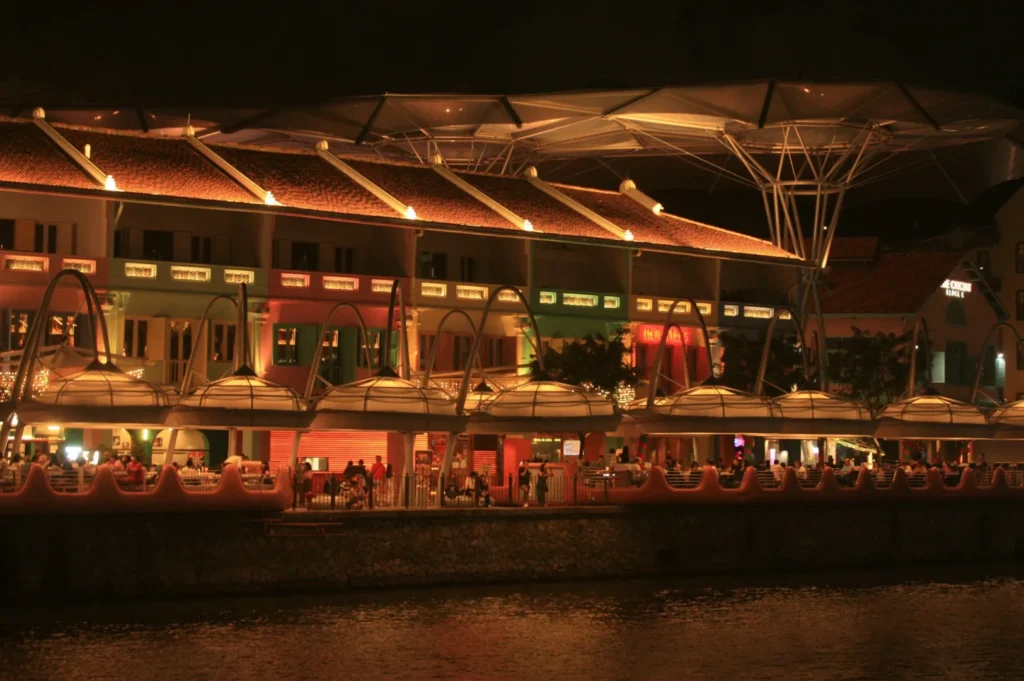 A vibrant waterfront scene at night, featuring a colorful building with bright lights and a large canopy roof, reflecting warmly off the water.