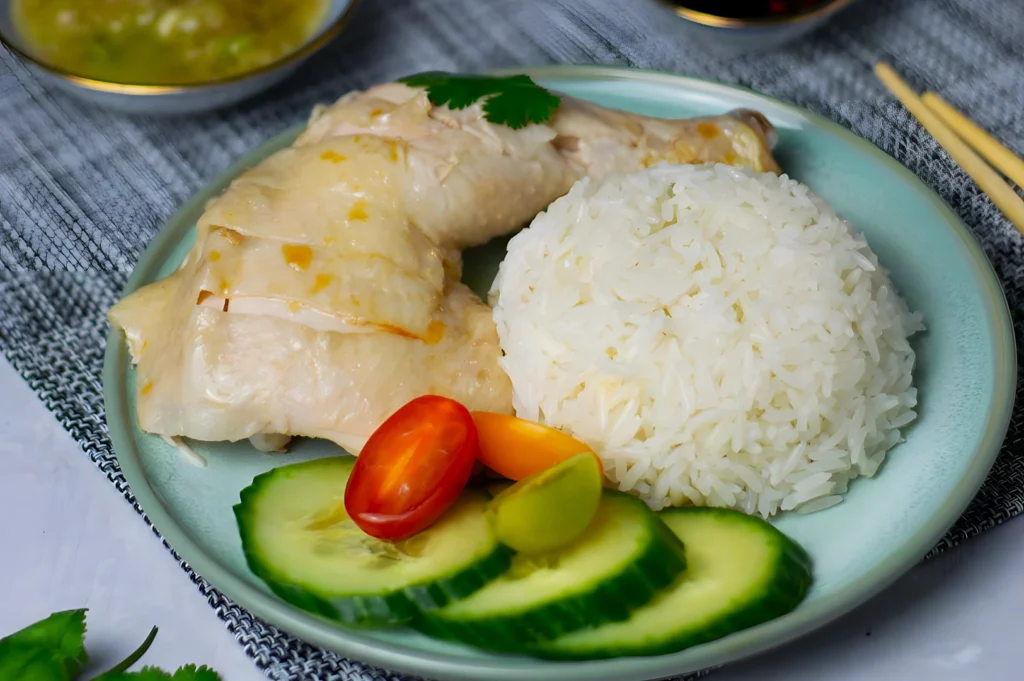 Plate featuring Hainanese chicken rice with tender chicken, a mound of white rice, sliced cucumbers, cherry tomatoes, and a sprig of cilantro.