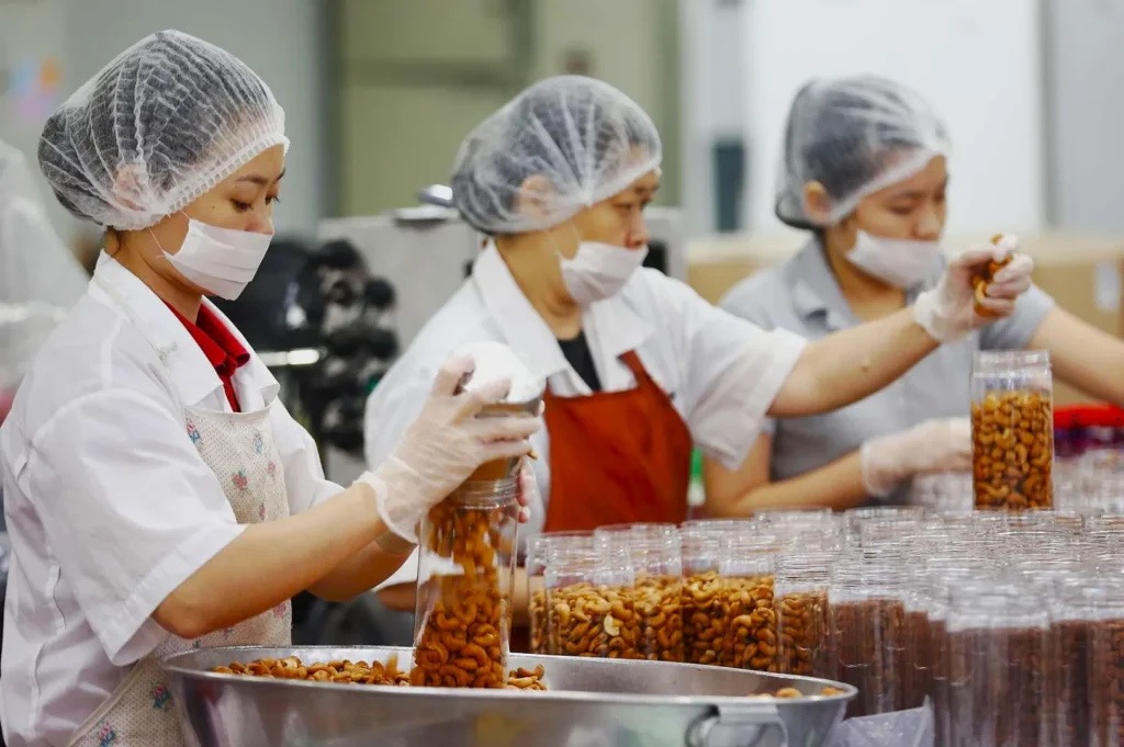 Three workers wearing hair nets and masks are packing cashews into clear containers in a factory setting. They appear focused and efficient.