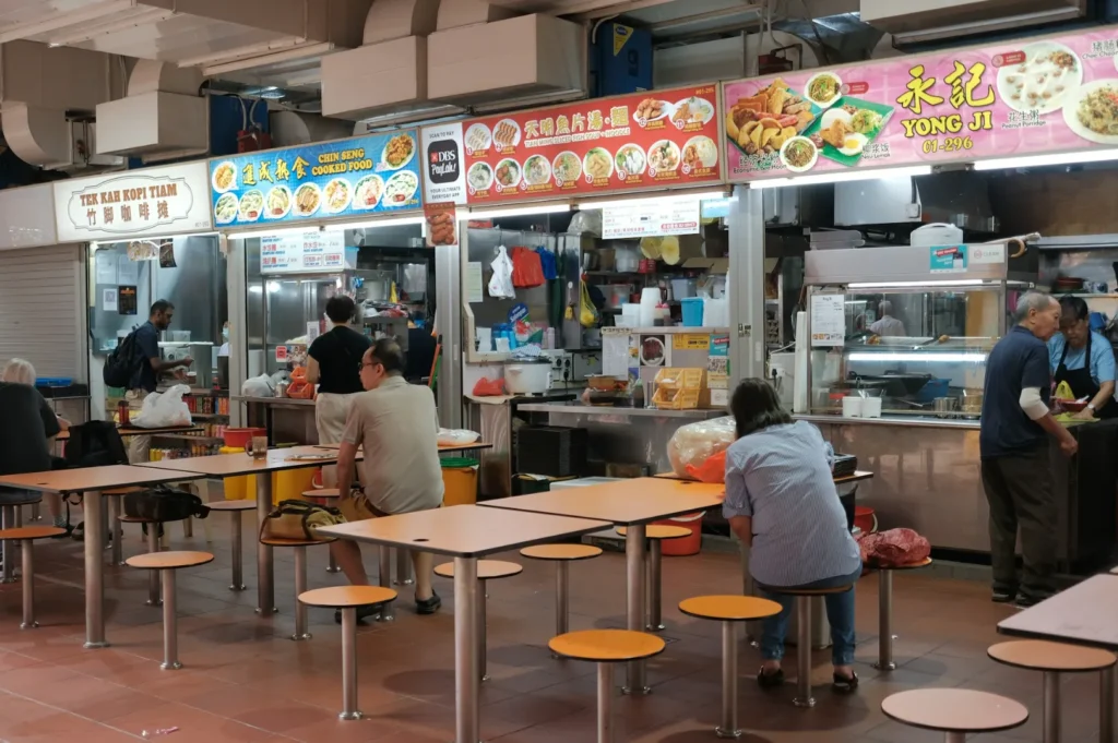 Indoor food court scene with several small food stalls and bright signs. Few people are seated at tables with orange stools, giving the place a casual, local ambiance.