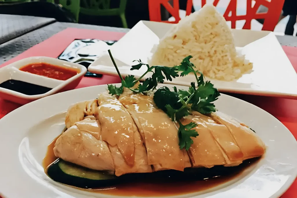 A plate of Hainanese chicken rice with tender, glazed chicken garnished with cilantro. Steamed rice in the background, with soy and chili sauces beside.