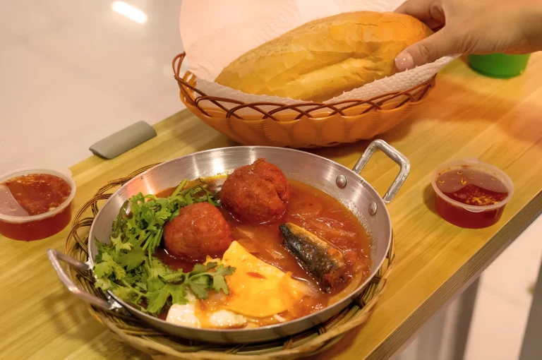 A Vietnamese dish with meatballs, herbs, cheese, and fish in tomato sauce is served in a pan. A hand reaches for a baguette. Two sauce cups are nearby.
