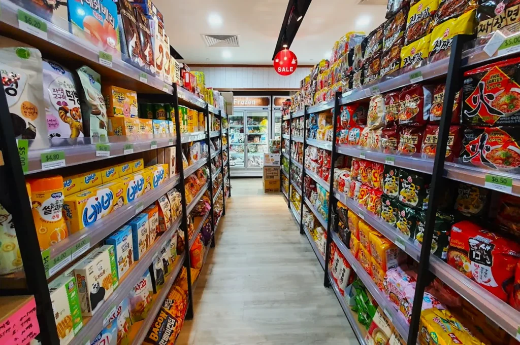 A brightly lit Asian grocery aisle with shelves full of colorful packaging. Instant noodles and snacks line both sides. A red "Sale" sign hangs overhead.