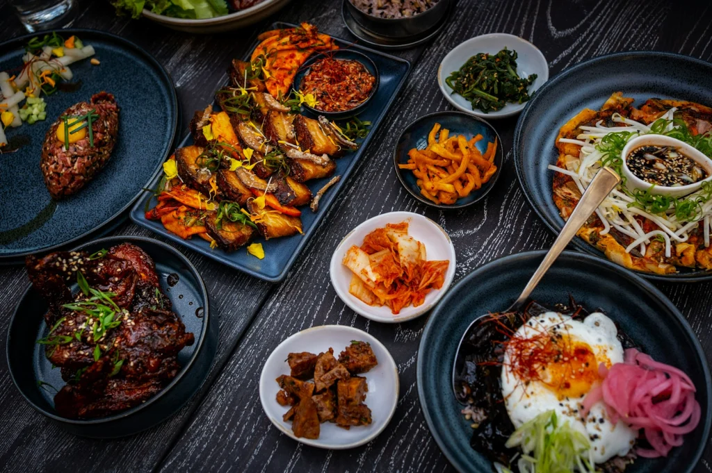 A variety of Korean dishes on a dark table, including kimchi, grilled meat, vegetables, and rice topped with a sunny-side-up egg, creating a vibrant, festive setting.