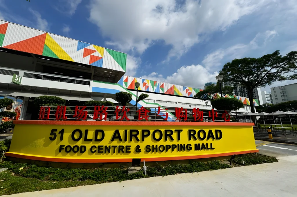 Exterior view of 51 Old Airport Road Food Centre & Shopping Mall in Singapore, featuring a large yellow and orange sign with the venue name in English and Chinese (旧机场路饮食与购物中心). The building behind displays a colorful geometric mural in red, green, yellow, and blue. Lush trees and a partly cloudy blue sky are visible in the background.