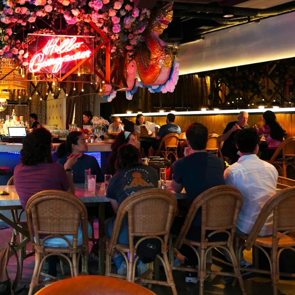 Dimly lit bar with patrons seated on wicker chairs around tables. A neon sign reads "Hello Gorgeous" amid hanging flowers, creating a cozy ambiance.