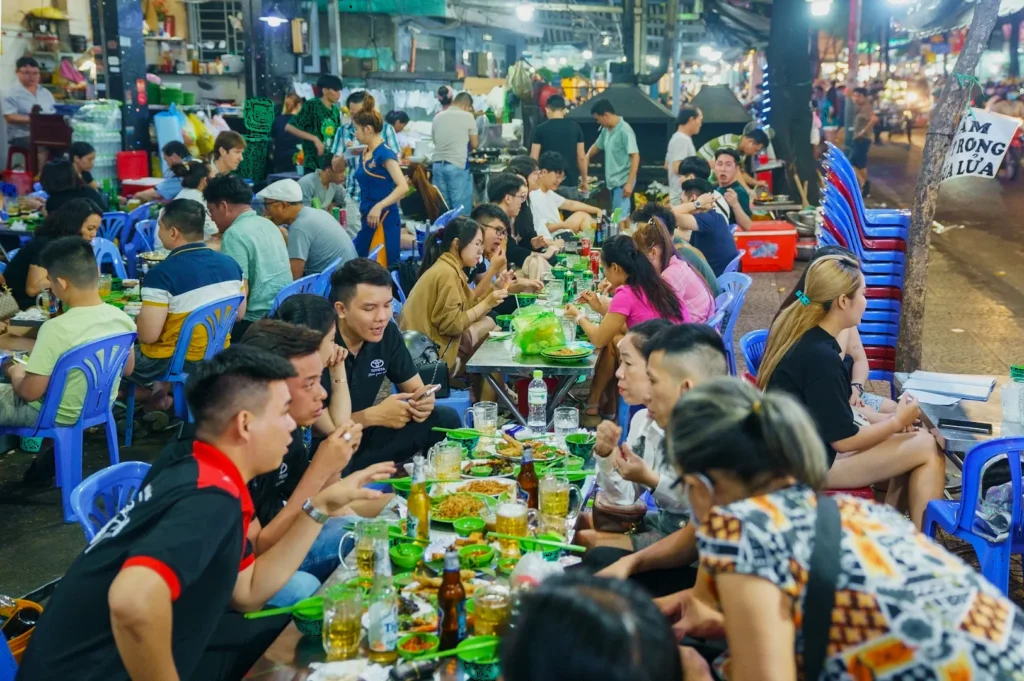 A bustling outdoor food market at night, crowded with people sitting at tables enjoying meals. The atmosphere is lively and social, with vibrant colors.