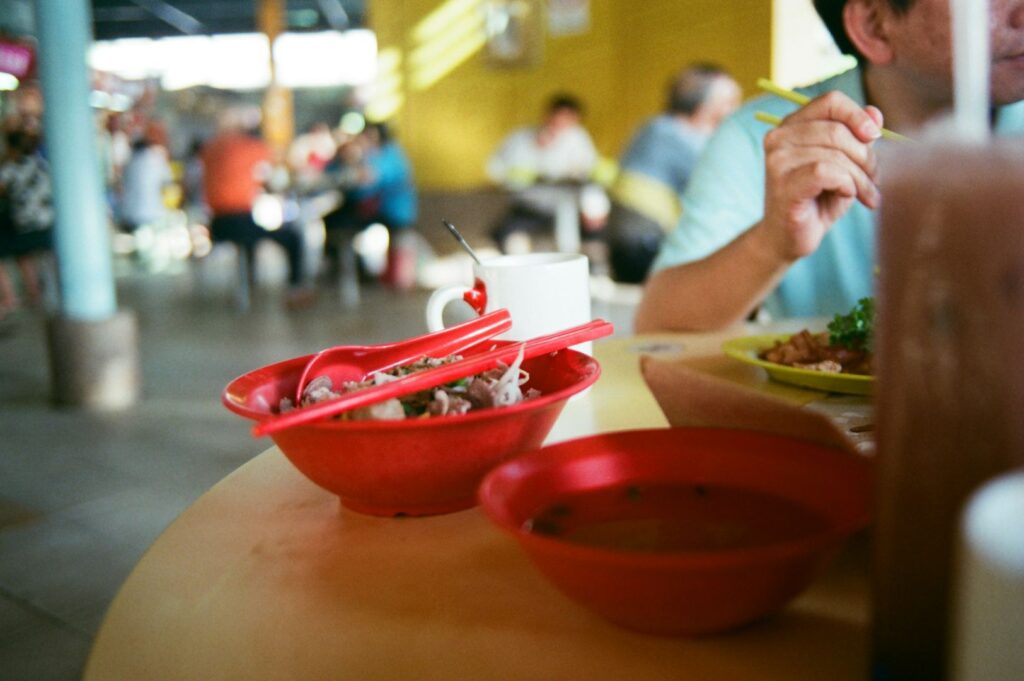 A bustling food court scene with a focus on a red bowl of noodles and chopsticks. A man eats nearby, with other diners blurred in the background. Casual and lively mood.