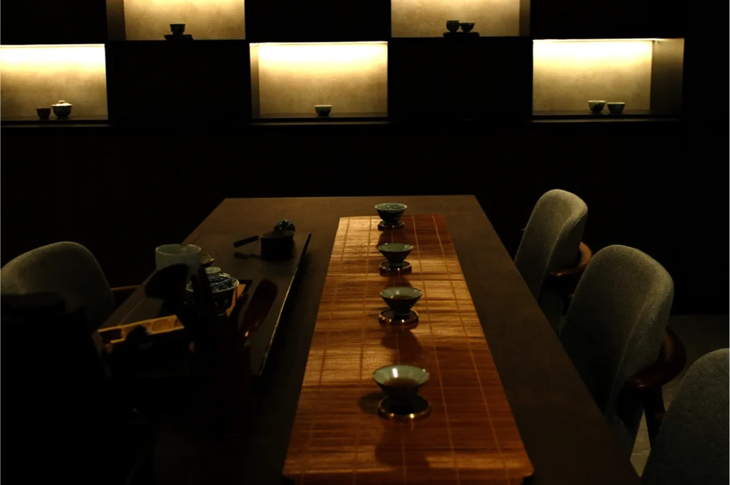 Dimly lit, elegant tea room with a long wooden table, bamboo runner, teacups in a row, and softly illuminated shelves displaying ceramics, evoking calm.