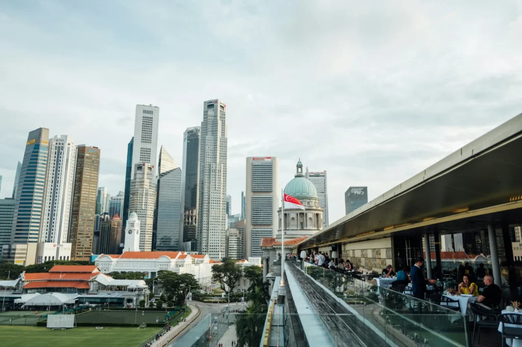Singapore skyline with modern skyscrapers, historic dome with a flag, and a terrace with diners under a cloudy sky. Urban and lively atmosphere.