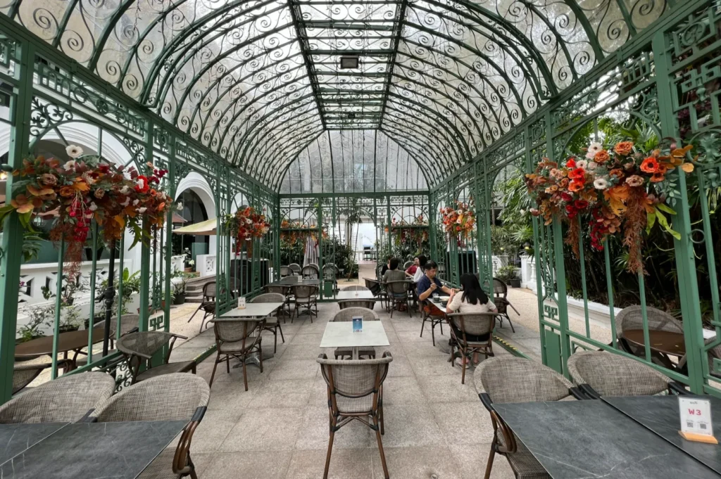 Glass-roofed courtyard cafe with ornate green metal framework, colorful floral arrangements, and several people seated at gray tables, creating a relaxed atmosphere.
