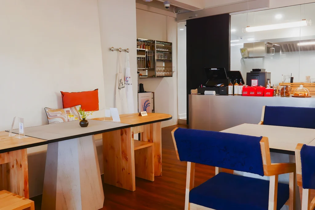 Modern cafe interior with wooden tables, blue cushioned chairs, and a counter with pastries. Soft lighting and minimal decor evoke a cozy atmosphere.