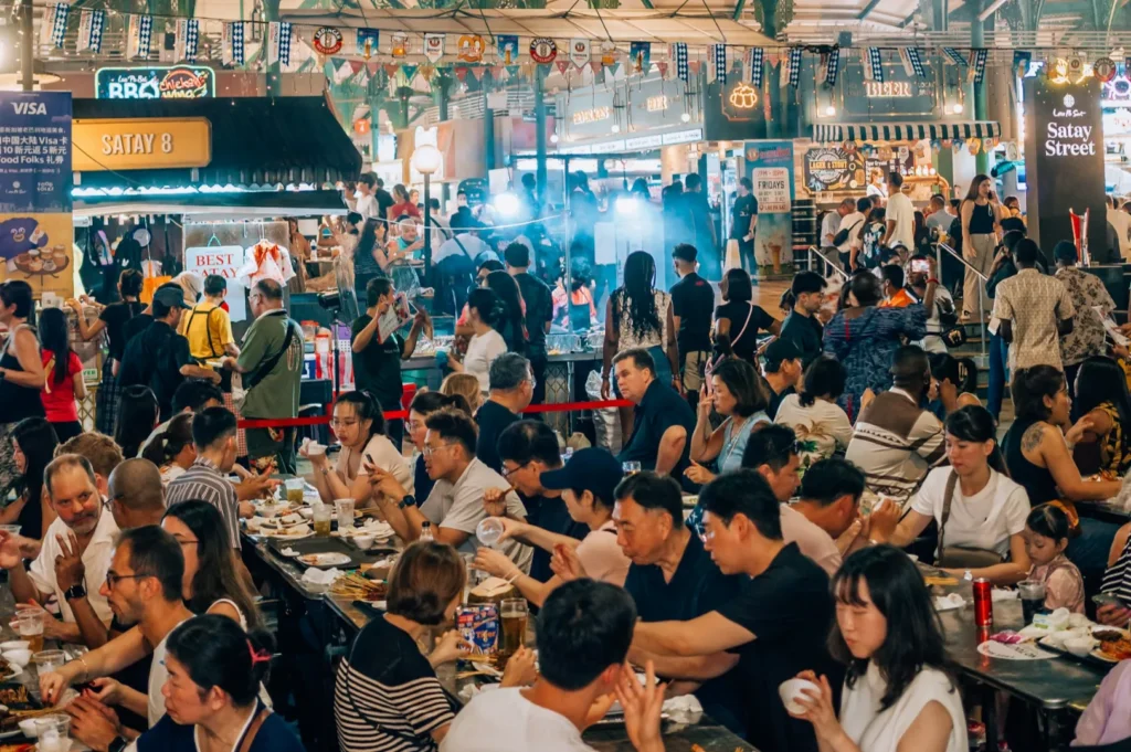 Bustling food market with people enjoying meals at communal tables. Colorful stalls, lively chatter, and smoke create a vibrant, festive atmosphere.