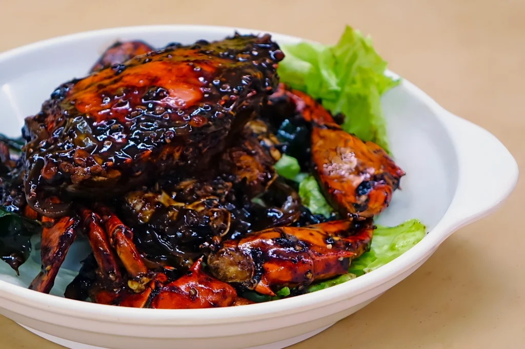 A dish of chili crab is served in a white bowl. The crab is coated in a rich, dark sauce and garnished with fresh lettuce, creating a savory appearance.