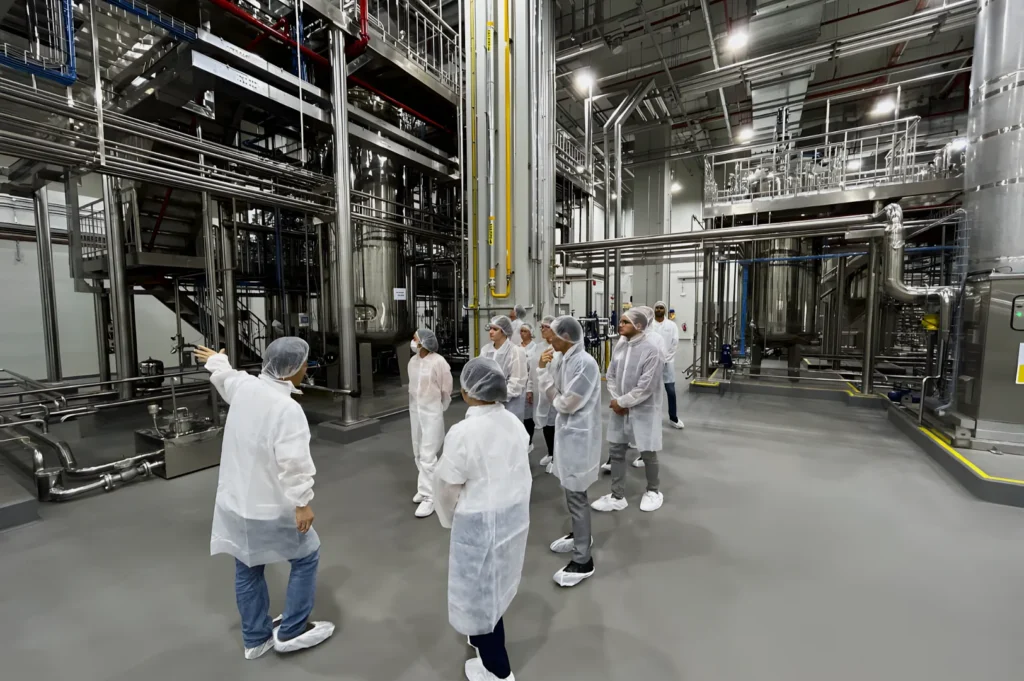 A group of people wearing protective clothing and hairnets are touring a modern industrial facility. One person gestures at machinery, explaining its function.