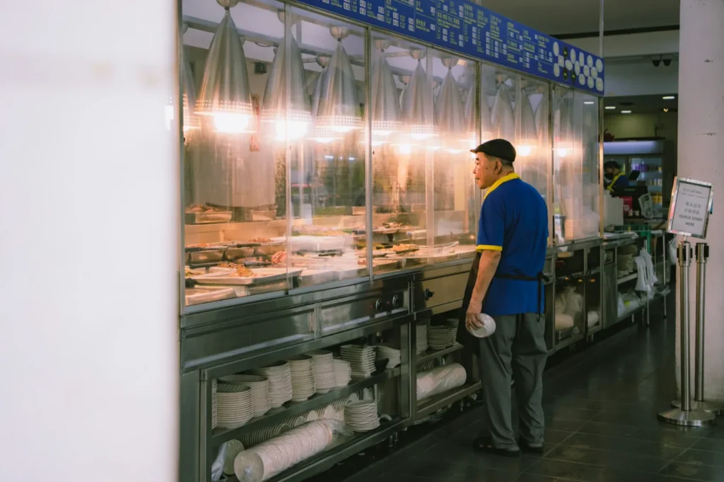 A man in a blue shirt and cap stands by a warmly lit food counter, holding a plate. Stacks of dishes are visible below, creating a cozy, inviting atmosphere.