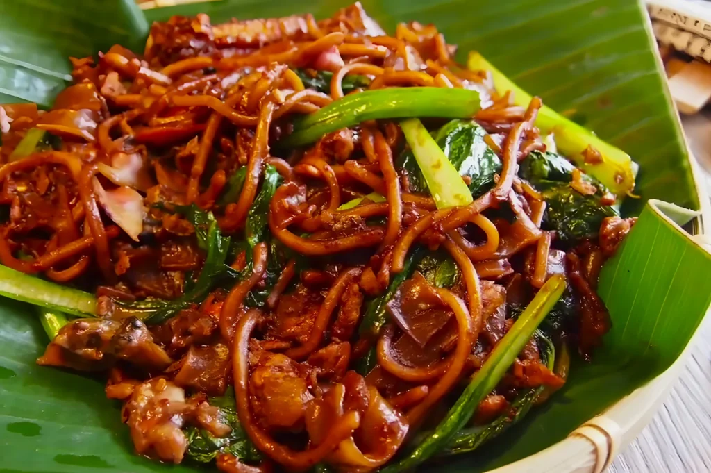 A close-up of stir-fried noodles with vegetables on a banana leaf. The dish includes green leafy vegetables and has a glossy, appetizing appearance.