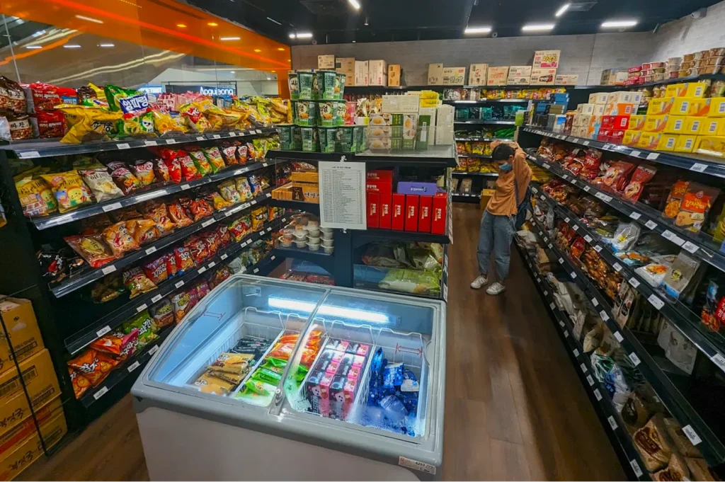 A small, brightly lit grocery store aisle filled with colorful snacks on shelves. A freezer displays frozen treats. A person shops, appearing thoughtful.
