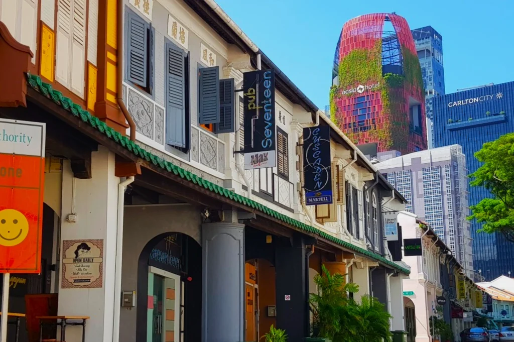 Historic shophouses with colorful facades and signs line a vibrant street. Modern skyscrapers, including a green-clad tower, rise in the background.