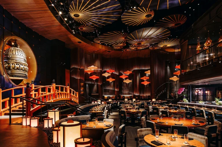 Luxurious Asian-themed restaurant interior featuring circular tables, elevated seating, warm lighting, intricate ceiling decor, and a large ornamental bell.