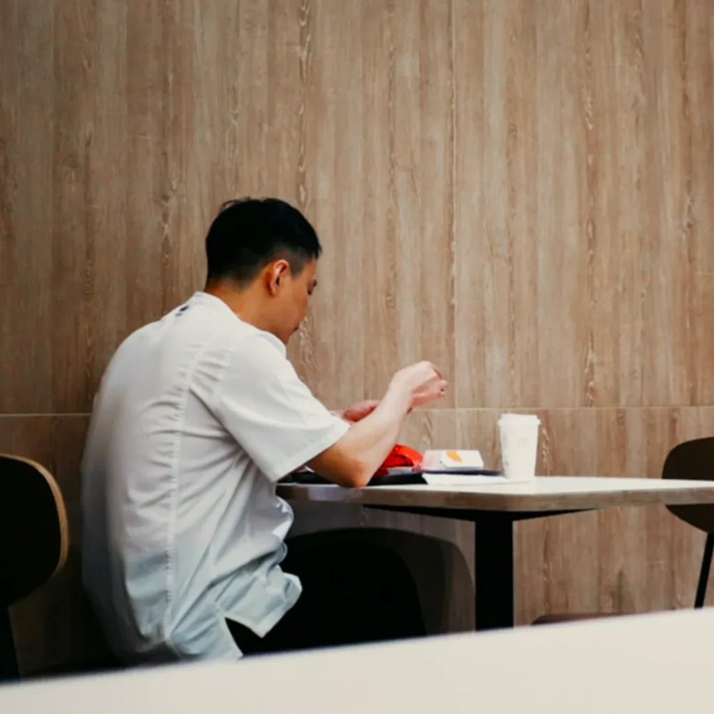 A person in a white shirt sits alone at a small table on a wooden panel wall background, eating fast food with a coffee cup beside them, conveying a calm and solitary atmosphere.