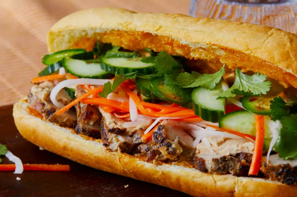 Close-up of a bánh mì sandwich with grilled meat, pickled carrots, daikon, cucumber slices, and cilantro in a crusty baguette on a wooden board.