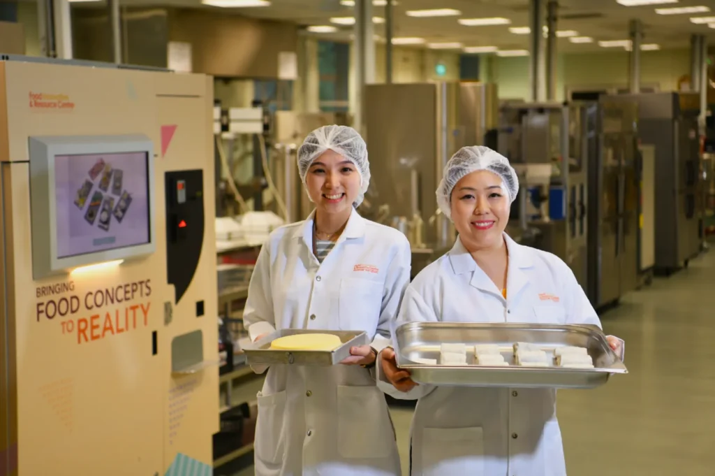 Two smiling women in lab coats and hairnets hold trays of food in a bright, industrial kitchen. A sign reads "Bringing Food Concepts to Reality."