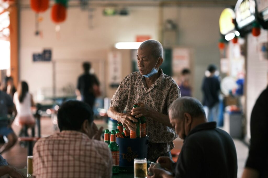 Elderly man with a face mask on his chin serves bottled drinks at a bustling, warmly-lit food court, creating a friendly, communal atmosphere.