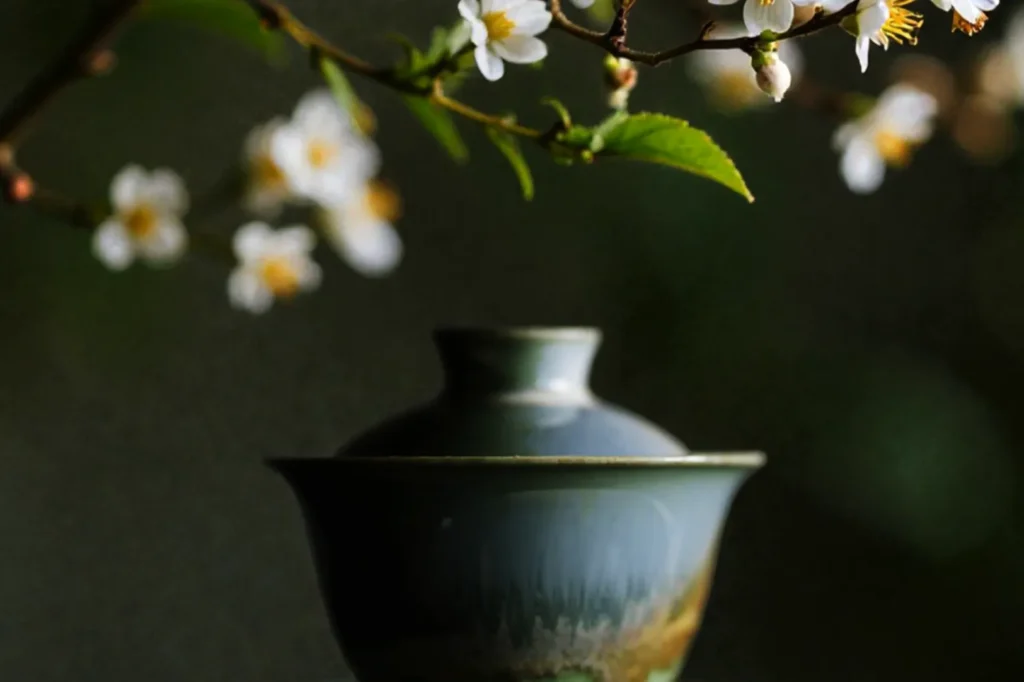 A ceramic cup with a lid sits beneath a branch of white cherry blossoms. The scene conveys tranquility and harmony with a soft-focus background.