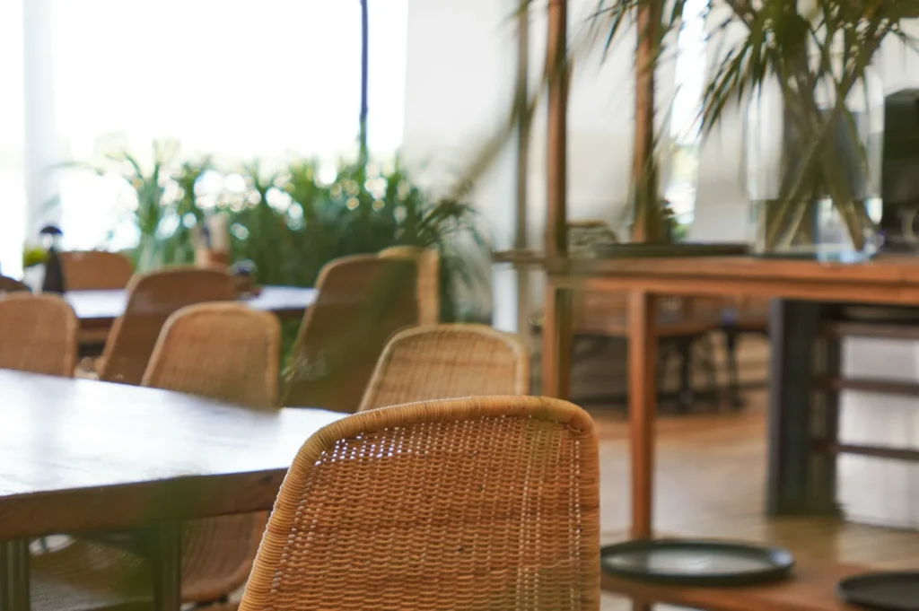 Cozy cafe interior with wicker chairs and wooden tables, surrounded by lush green plants. Sunlight filters through large windows, creating a relaxed atmosphere.