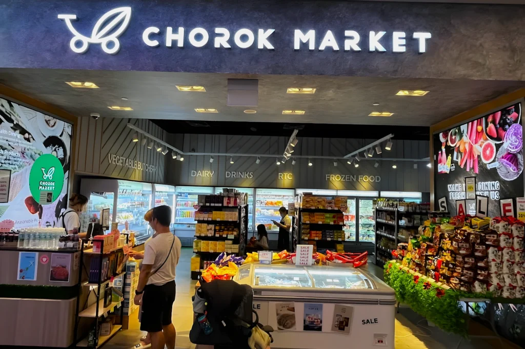 A brightly lit grocery store entrance labeled "Chorok Market" features sections for dairy, drinks, and frozen food. Shoppers browse snacks and sales displays.