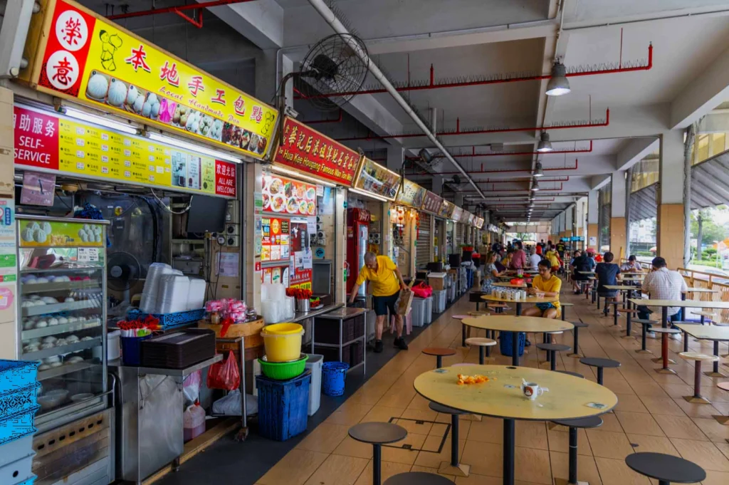 A bustling food court with various food stalls and colorful signs. Diners sit at round tables, creating a vibrant and lively atmosphere.