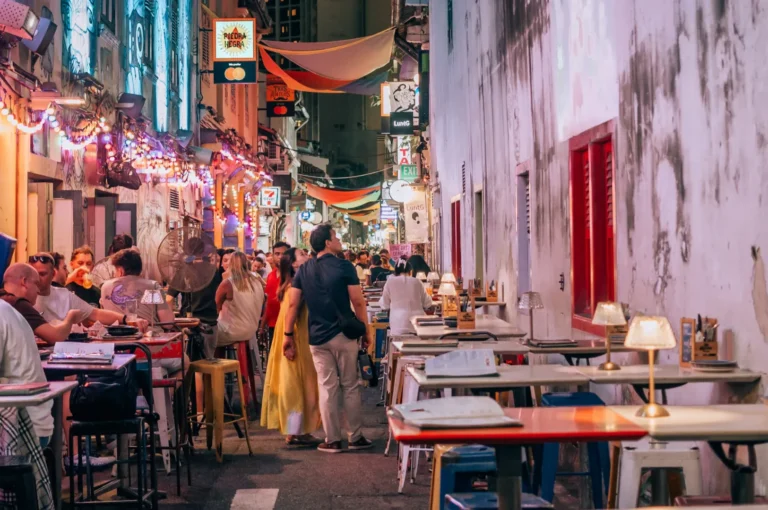A vibrant alley brims with diners at outdoor tables under colorful lights and signs. The lively atmosphere evokes warmth and social joy, with people chatting and dining.