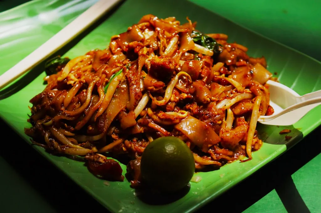 A plate of Pad Thai with noodles, bean sprouts, and chicken, served on a green dish. A lime wedge and a spoon are placed beside the dish.