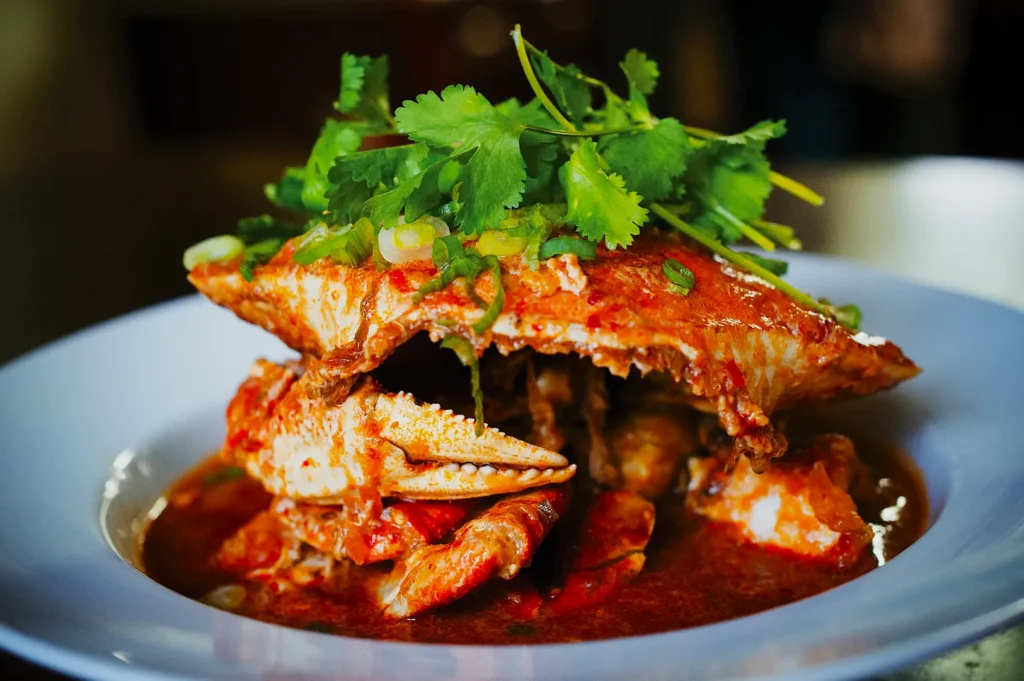 A dish of cooked crab in rich red sauce, garnished with fresh cilantro, is served in a white bowl. The presentation is vibrant and appetizing.