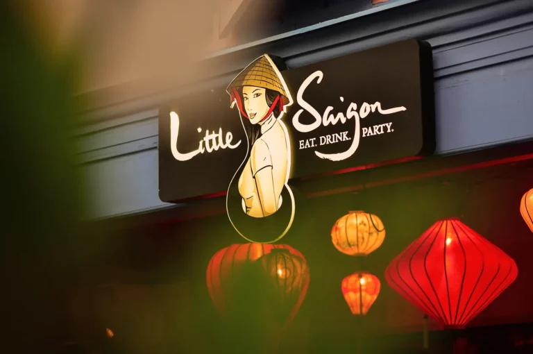 Sign for "Little Saigon" with a stylized illustration of a woman in a conical hat. Surrounded by warm, red lanterns creating a festive, inviting atmosphere.