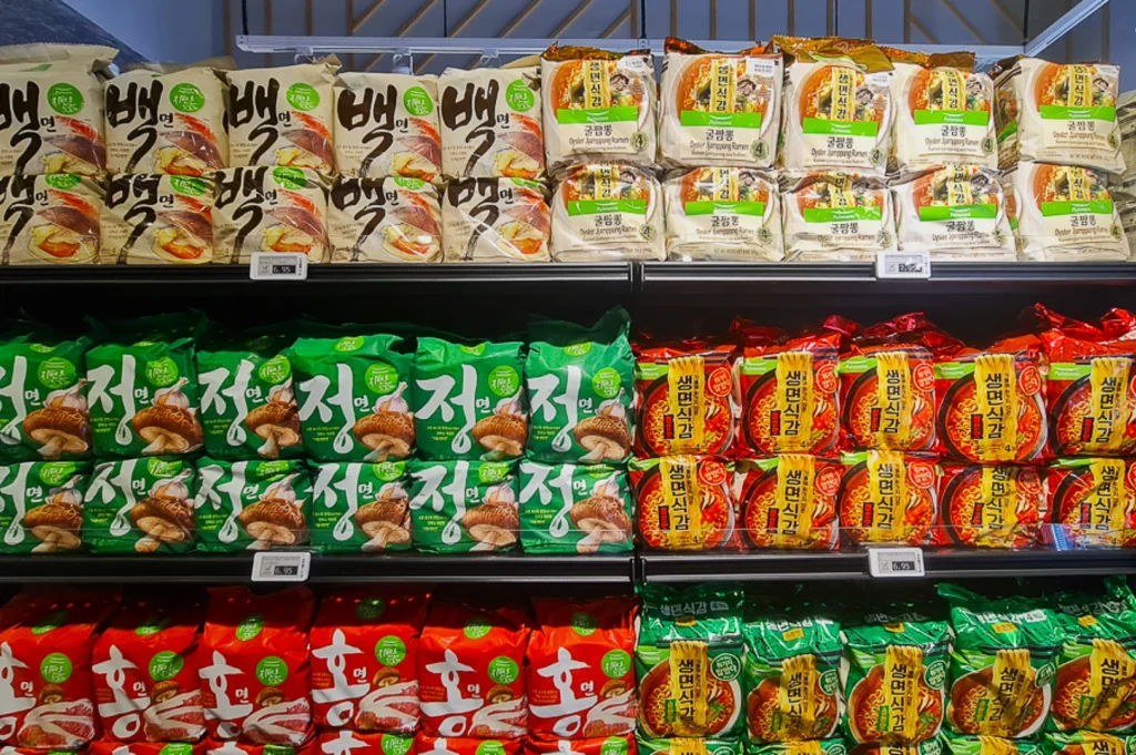 Grocery shelves display assorted Korean instant noodles in colorful packaging. Top shelf shows white packs, middle green, bottom red, evoking a diverse, vibrant selection.