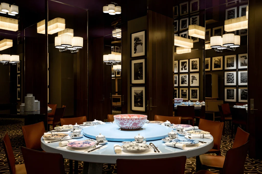 Elegant dining room with a round table set for eight. A large decorative bowl is central, surrounded by floral china. Warm lighting and framed art create a sophisticated ambiance.