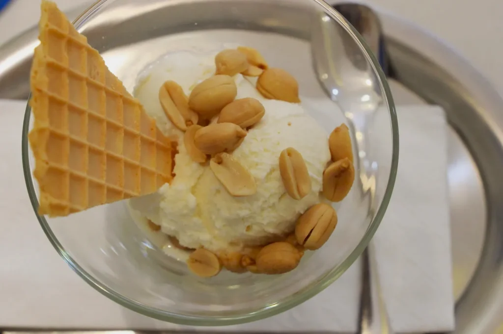 Vanilla ice cream in a glass bowl topped with peanuts and a waffle triangle. A spoon rests beside the bowl on a metallic tray with a napkin.