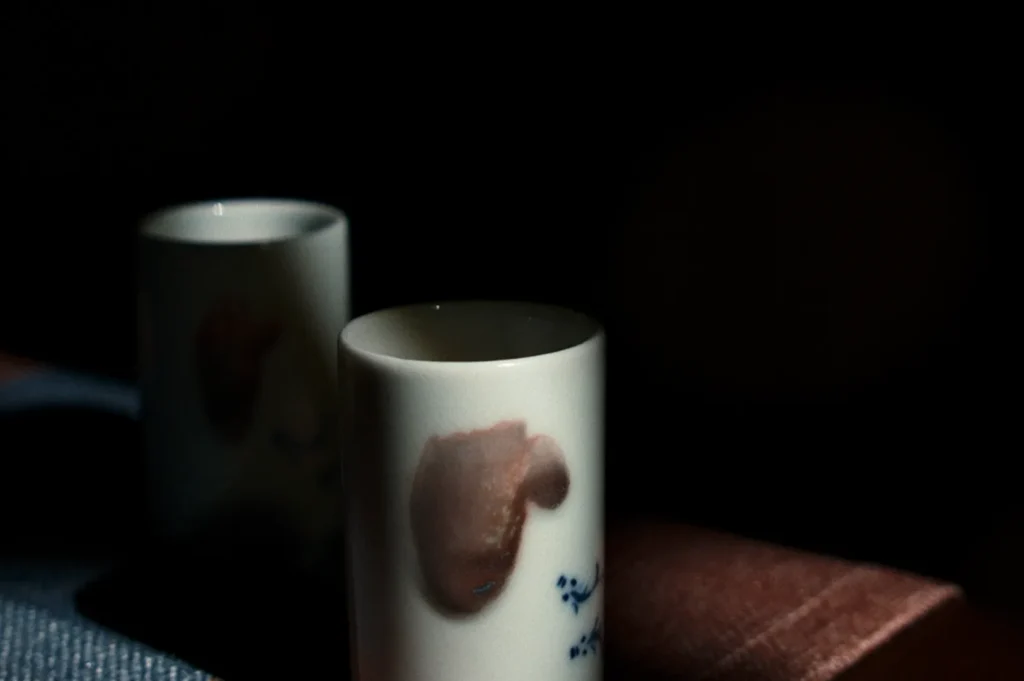 Two ceramic cups with abstract patterns are partially lit, creating a dramatic contrast against the dark background. A subtle, serene mood is conveyed.