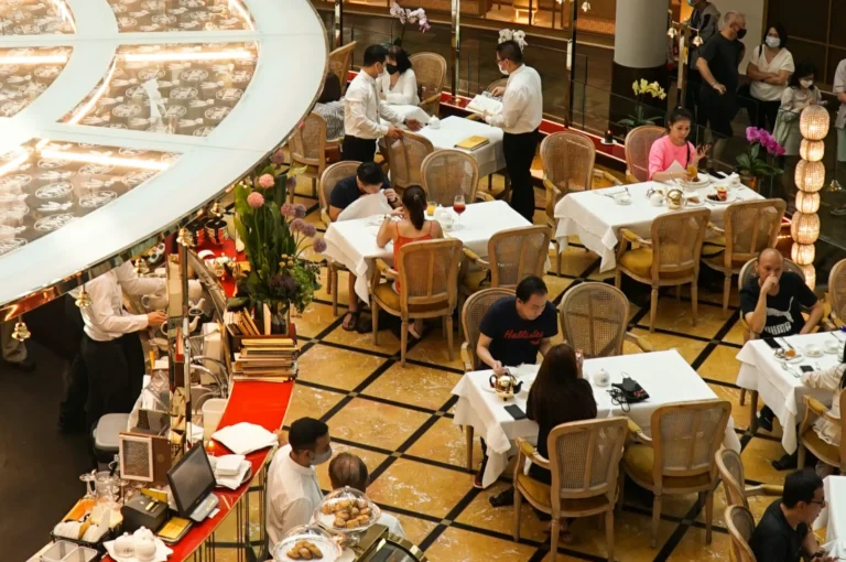 A bustling restaurant scene with elegantly set tables, patrons dining, and servers in white uniforms attending to guests. The atmosphere is lively and upscale.