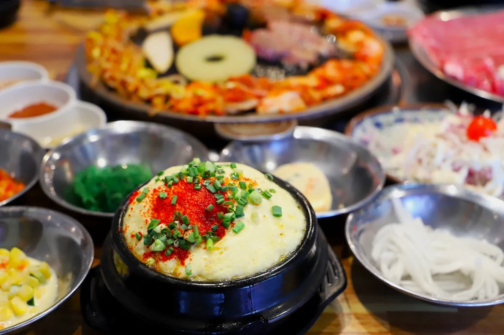 A Korean table spread featuring a hot stone pot with steamed egg garnished with spring onions and red seasoning, surrounded by vibrant side dishes.
