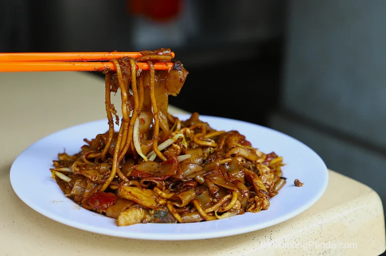 A plate of char kway teow, featuring stir-fried flat rice noodles with bean sprouts and meat. Orange chopsticks hold a portion above, emphasizing its glossy texture.
